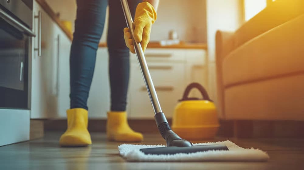 Person mopping kitchen floor in yellow gloves and boots with a vacuum cleaner nearby.
